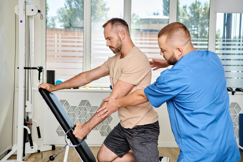 experienced physiotherapist helping man in sportswear during recovery training on exercise machine